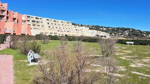 Un immeuble résidentiel à plusieurs étages doté de balcons, une zone verdoyante avec un banc et un ciel bleu dégagé.