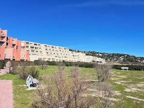 A multi-story residential building with balconies, a grassy area with a bench, and a clear blue sky. - Cosy studio with sea & garden view - swimming pool (Sète)