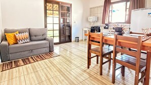 A compact living space with a sofa, dining table, chairs, a kitchen area, and a sliding glass door. - Chalés Caminho das Pedras S. B. Sapucaí (São Bento do Sapucaí)