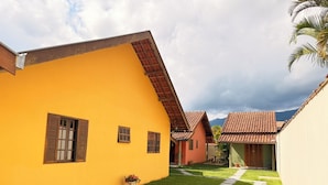 A yellow building with a sloped roof, a small garden area with a pathway, and a mountain in the background. - Chalés Caminho das Pedras S. B. Sapucaí (São Bento do Sapucaí)