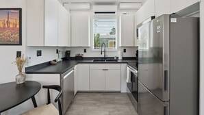 Une cuisine moderne avec des meubles blancs, un réfrigérateur en acier inoxydable, un plan de travail noir et une fenêtre équipée de stores.