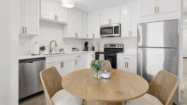 Une cuisine moderne avec des meubles blancs, des appareils en acier inoxydable et une table ronde en bois, accompagnée de chaises en osier.