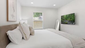 Une chambre à coucher avec un lit, des oreillers, une fenêtre avec des stores, un tableau et une vue sur la verdure.