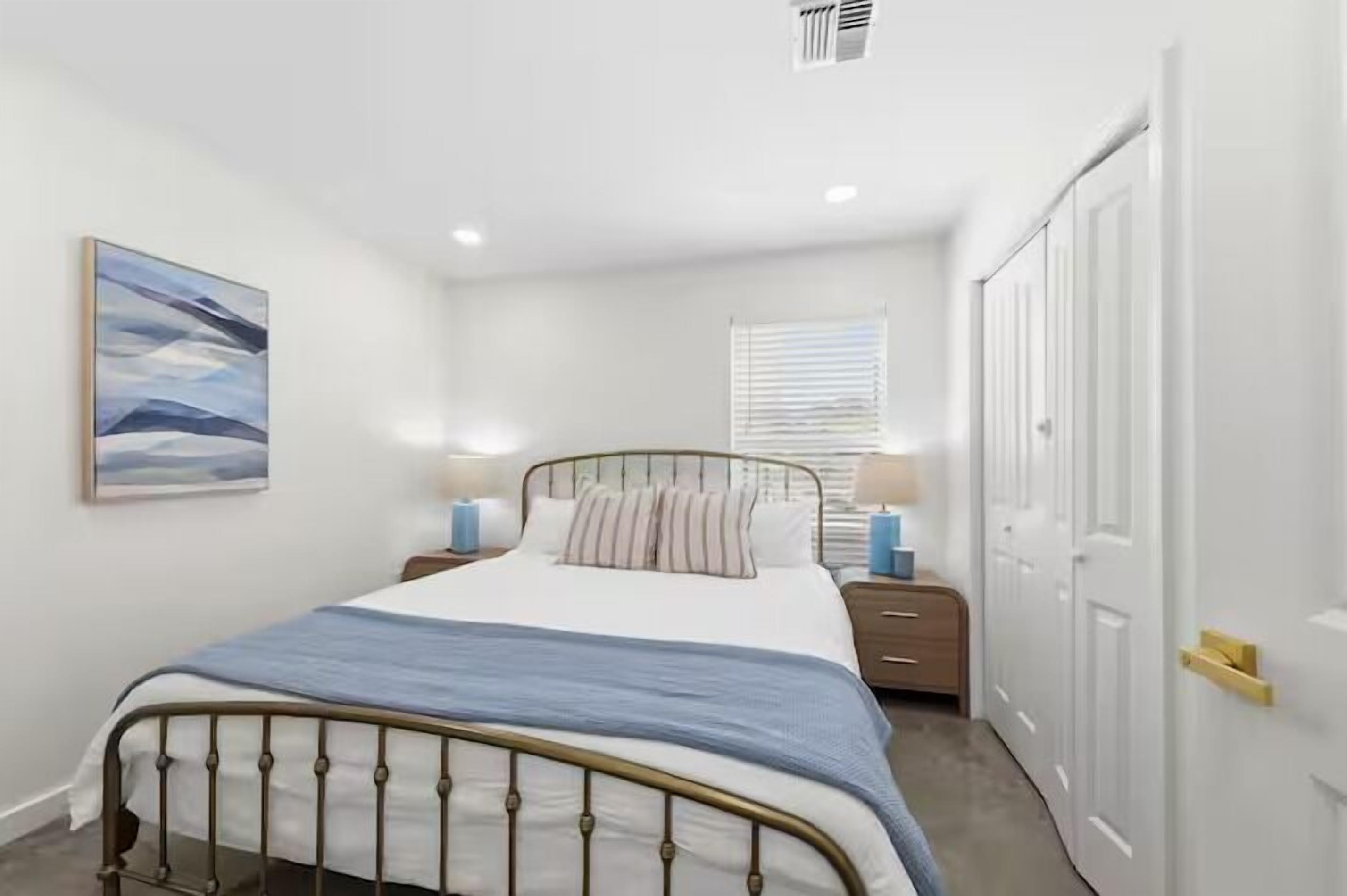 A bedroom with a bed, bedside tables, a window with blinds, a door, and a painting on the wall.