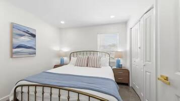 A bedroom with a bed, bedside tables, a window with blinds, a door, and a painting on the wall.