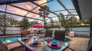 Et overdækket udendørs område med pool, spisebord, stole og en rød parasol.