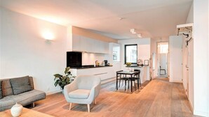 Un salon moderne comprenant un canapé, un fauteuil et une table basse, un coin cuisine avec des placards et un téléviseur, ainsi qu’un coin repas avec des chaises et une table.