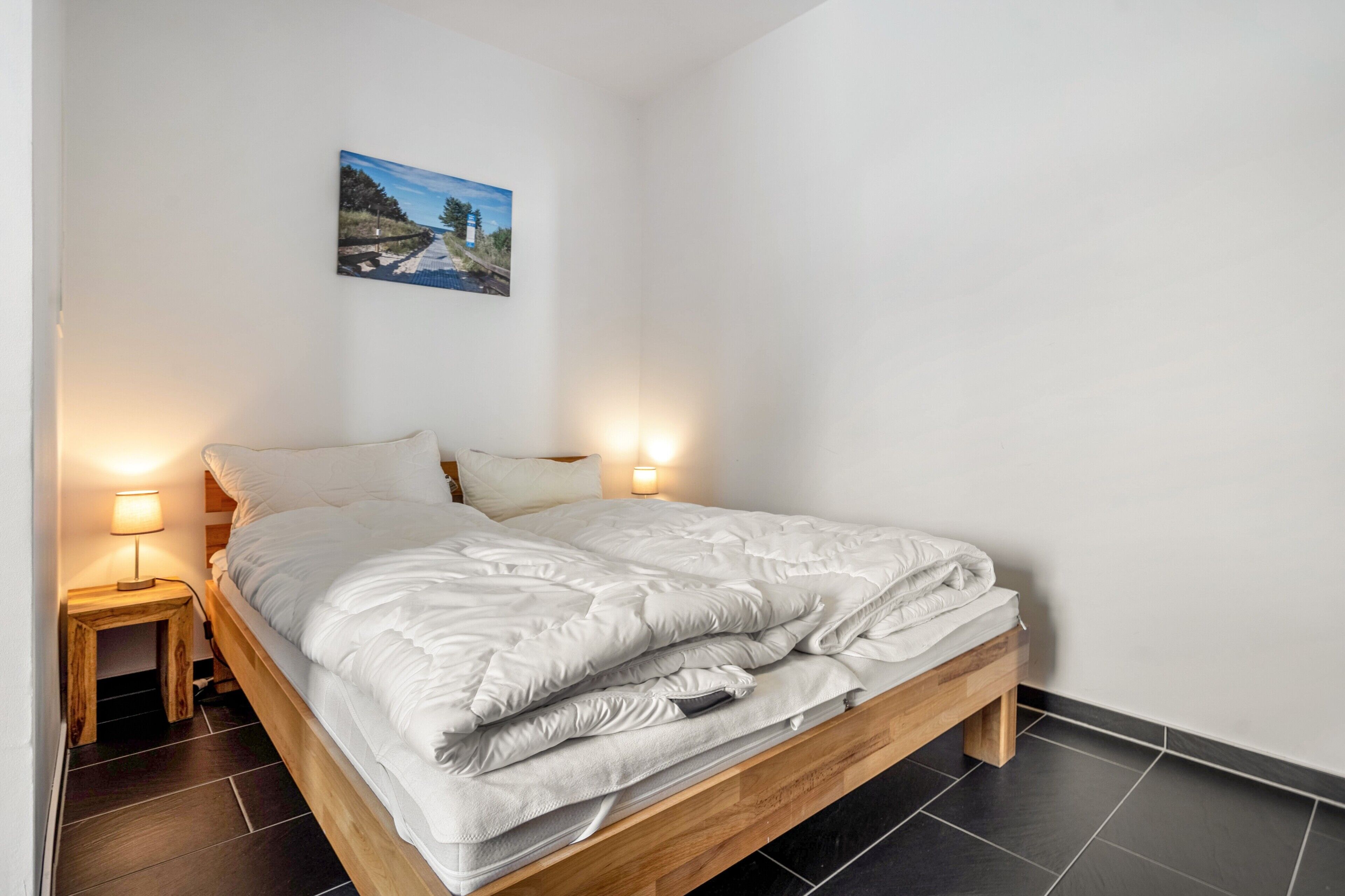Ein Einzelbett mit Holzrahmen, weiße Bettwäsche, zwei Nachttische mit Lampen und ein an der Wand hängendes Bild.