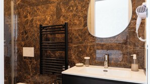 Un baño con pared de mármol, un toallero negro, un espejo redondo, un secador de pelo, un lavabo con grifo y dispensador de jabón.
