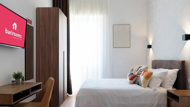 Habitación de hotel con cama, escritorio, silla, televisor y ventana con cortinas.
