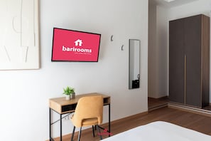 Moderni hotellihuone, jossa on puinen työpöytä, tuoli, ruukkukasvi, seinälle asennettu televisio, peili ja vaatekaappi.