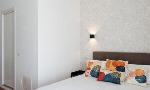 Un dormitorio con cama, almohadas, mesita de noche, lámpara de pared y una puerta.