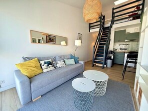 A modern living room with a grey sofa, a staircase, a kitchen, and a dining area. - 34 m² Studio with Private Parking in Rochelle (La Rochelle)