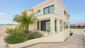 A modern two-story building with a balcony, a small garden with plants, and a view of the sea.