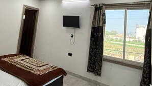 A bedroom with a bed, a flat-screen TV, a ceiling fan, a window with curtains, and a view of buildings outside. - Vidya Dham Vrindavan  (Vrindavan)