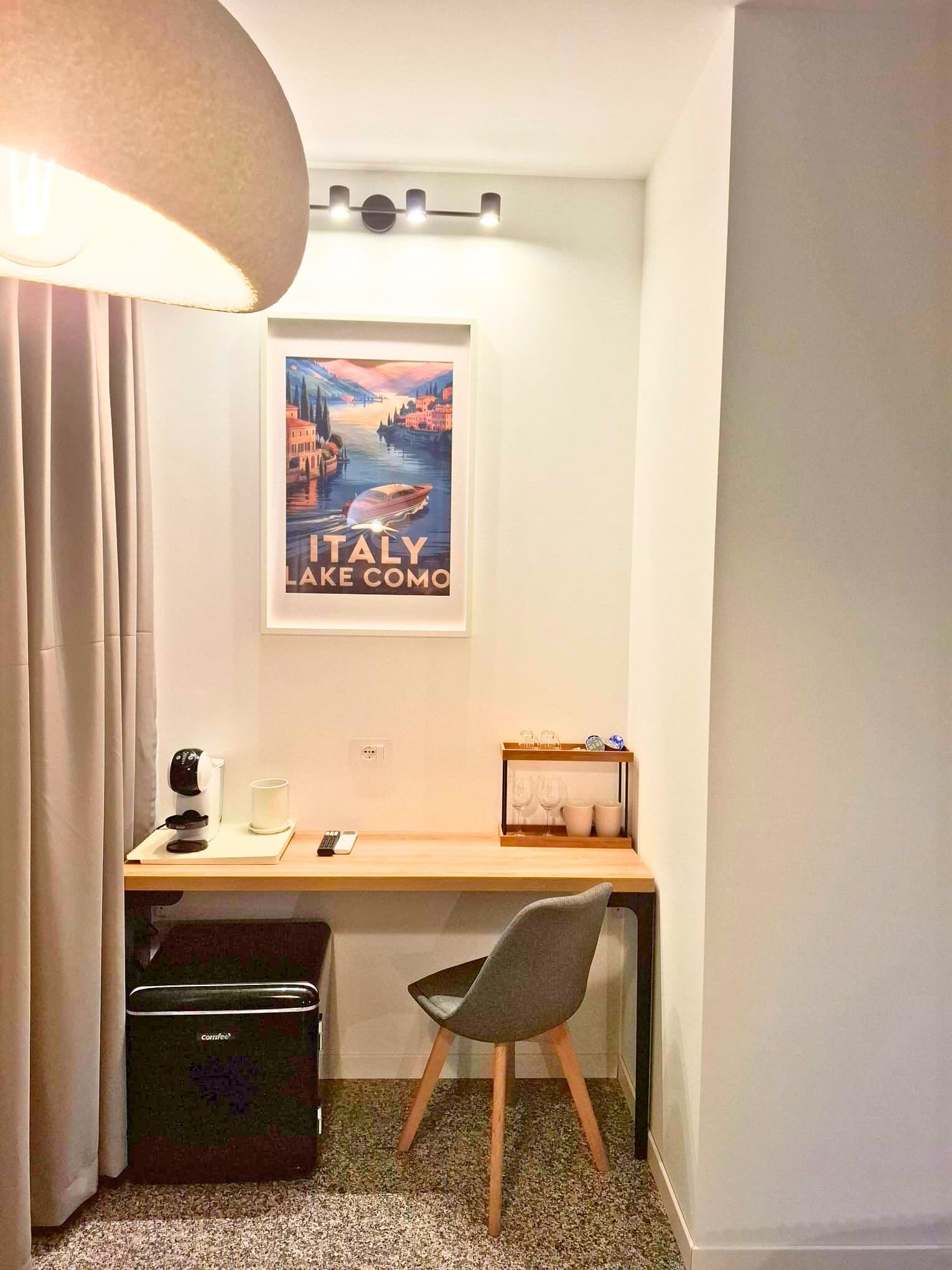 A small room with a desk, chair, coffee machine, and a framed picture of Italy.