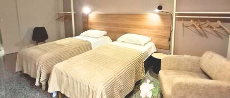 A hotel room with two beds, a round wooden table, a chair, a hanging light fixture, and a coat rack with hangers.