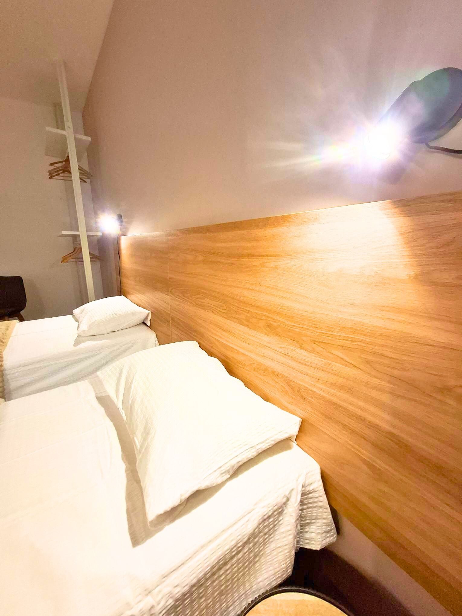 A hotel room with a wooden headboard, two beds with white linens, a white shelf unit, and a wall-mounted light fixture.