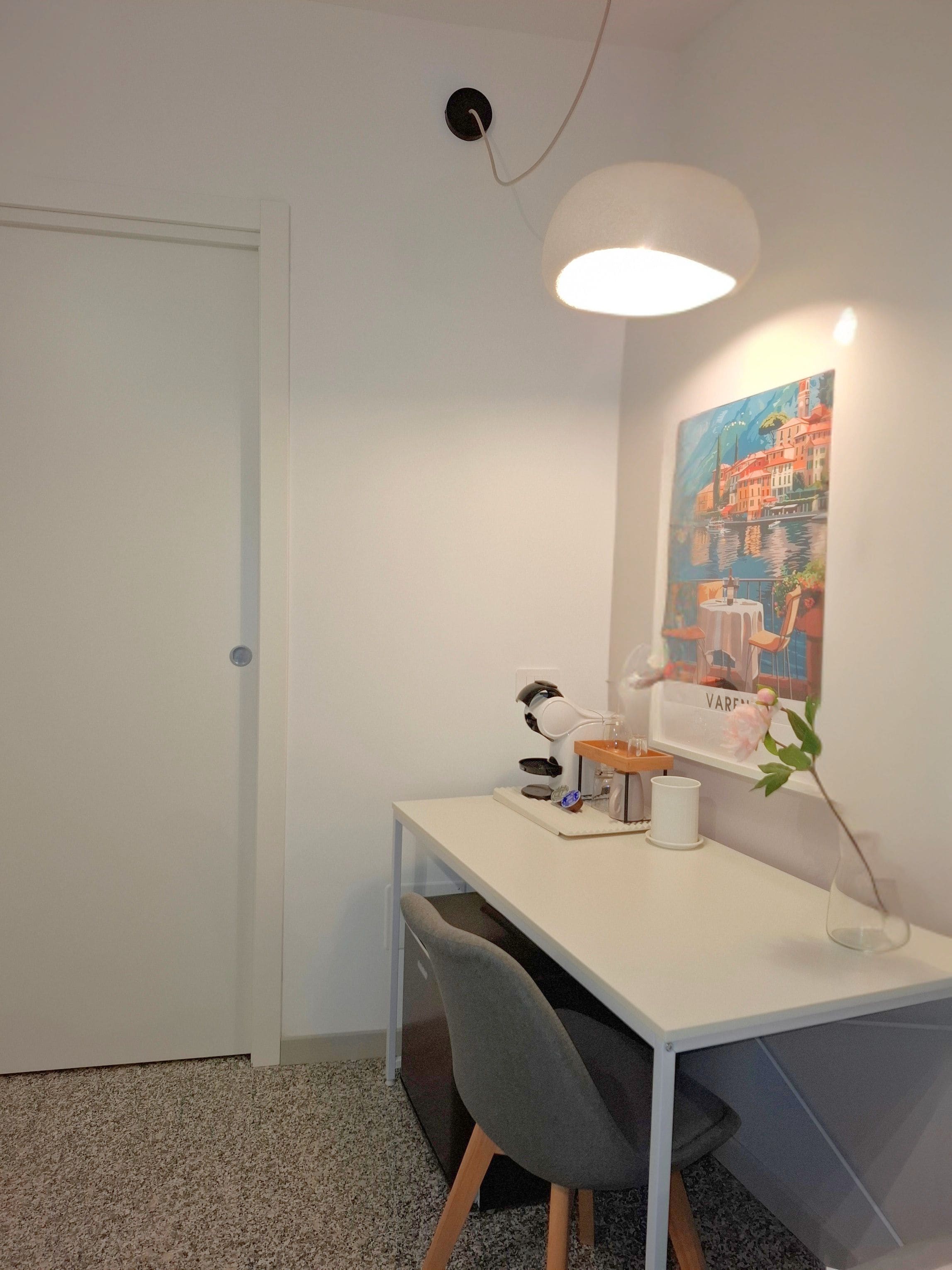 A white desk with a computer, a lamp, and a painting of a coastal scene.