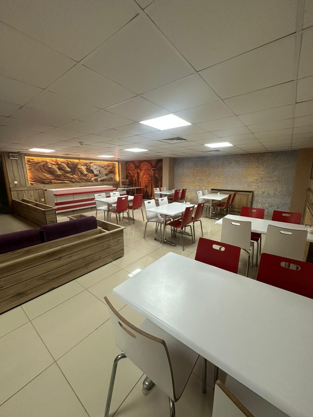 A restaurant with white tables and red chairs, a wooden bench area, and a mural on the wall.