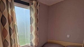 A small, single-room accommodation with two beds, a window with patterned curtains, and a plain wall.