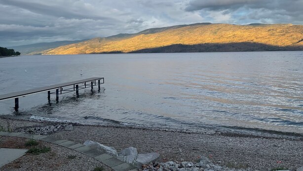 Ein ruhiger See mit einer hölzernen Mole, die sich ins Wasser erstreckt, umgeben von felsigem Gelände und in der Ferne Bergen unter einem bewölkten Himmel.