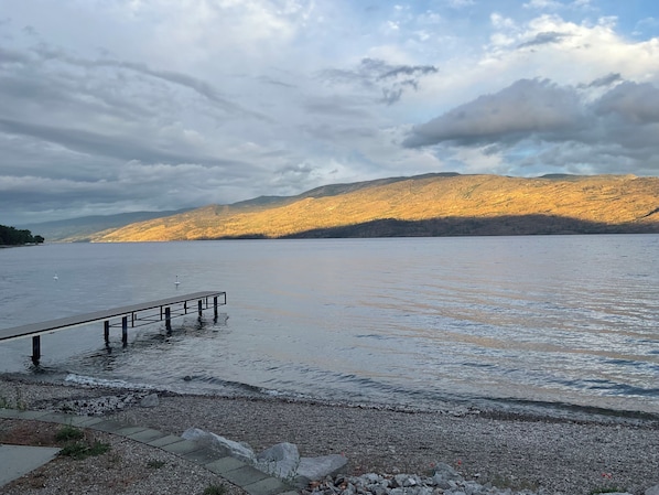 A calm lake with a wooden pier extending into the water, surrounded by rocky terrain and distant mountains under a cloudy sky.