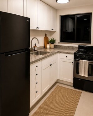 A kitchen with white cabinets, a black refrigerator, a stainless steel sink, a wooden cutting board, a potted plant, and a tiled backsplash. - 418 Luxury - Living 2 (Miami)