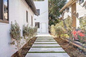 A narrow pathway leading to a white building with a door, flanked by small trees and a chain-link fence. - 418 Luxury - Living 2 (Miami)