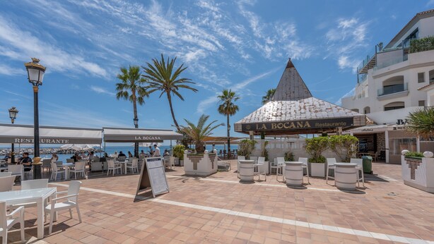 An outdoor dining area with tables and chairs, palm trees, and a building with 'LA BOCANA Restaurante' signage. - Holiday Home 'Enjoy Puerto Banus Next To Beach' with Sea View, Wi-Fi and Air Conditioning (Marbella)