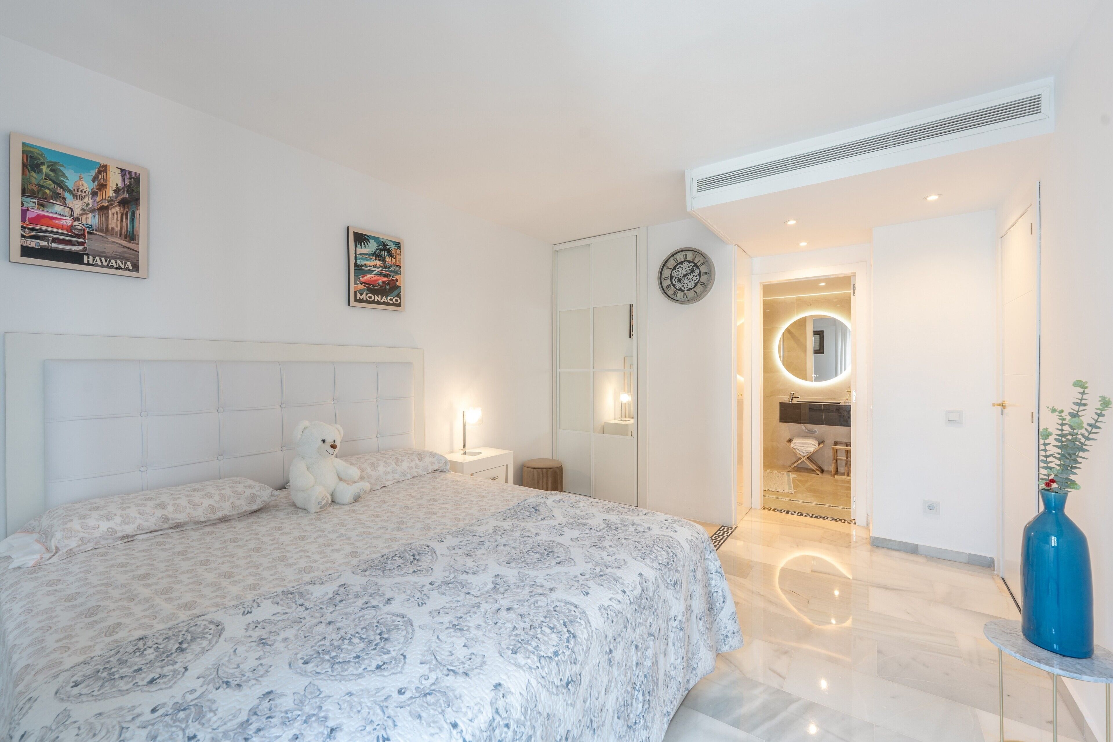 A bedroom with a bed, a teddy bear, a nightstand, a wall clock, a mirror, a bathroom with a sink, and a vase with a plant.