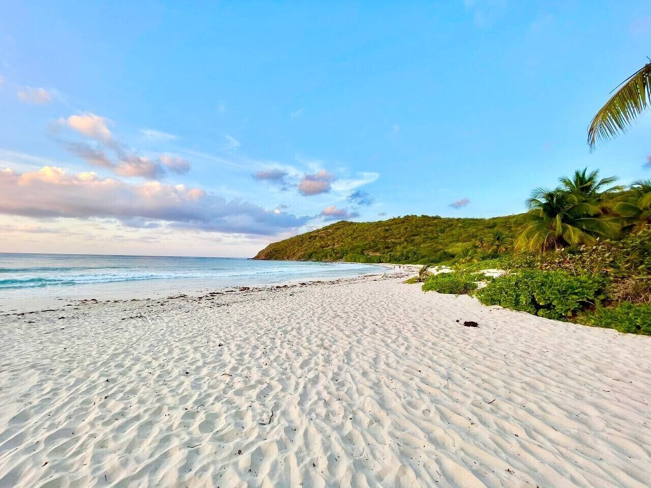 Una playa de arena blanca, cielo despejado y una colina lejana con vegetación.