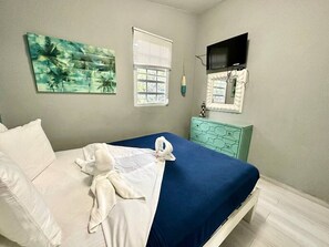 A bed with a blue bedspread, white pillows, and a white towel. There is a window with blinds, a painting on the wall, and a television mounted above the bed. - Villa 11 Flamenco Beachfront Villa (Flamenco)