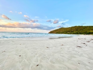 En strand med en sandstrand, vågor som slår in mot land och en grön kulle i bakgrunden.