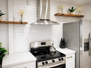 A modern kitchen with a stainless steel stove, a built-in oven, and a refrigerator. There are wooden shelves with decorative plants and a white tiled backsplash. - Beautiful 1 Bedroom Sun Lit Scottsdale Condo Welcome To Old Town Scottsdale (Scottsdale)