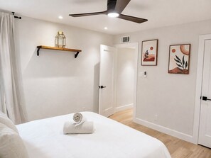 Ett hotellrum med en säng, en takfläkt, en hylla med en dekorativ prydnad och två inramade konstverk på väggen.
