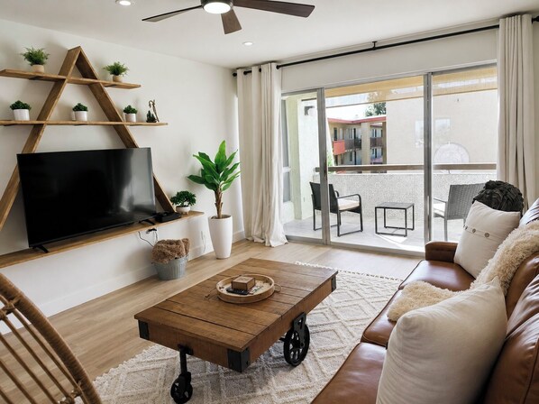 Un soggiorno con televisore, tavolino in legno, divano e un balcone con mobili da esterno.