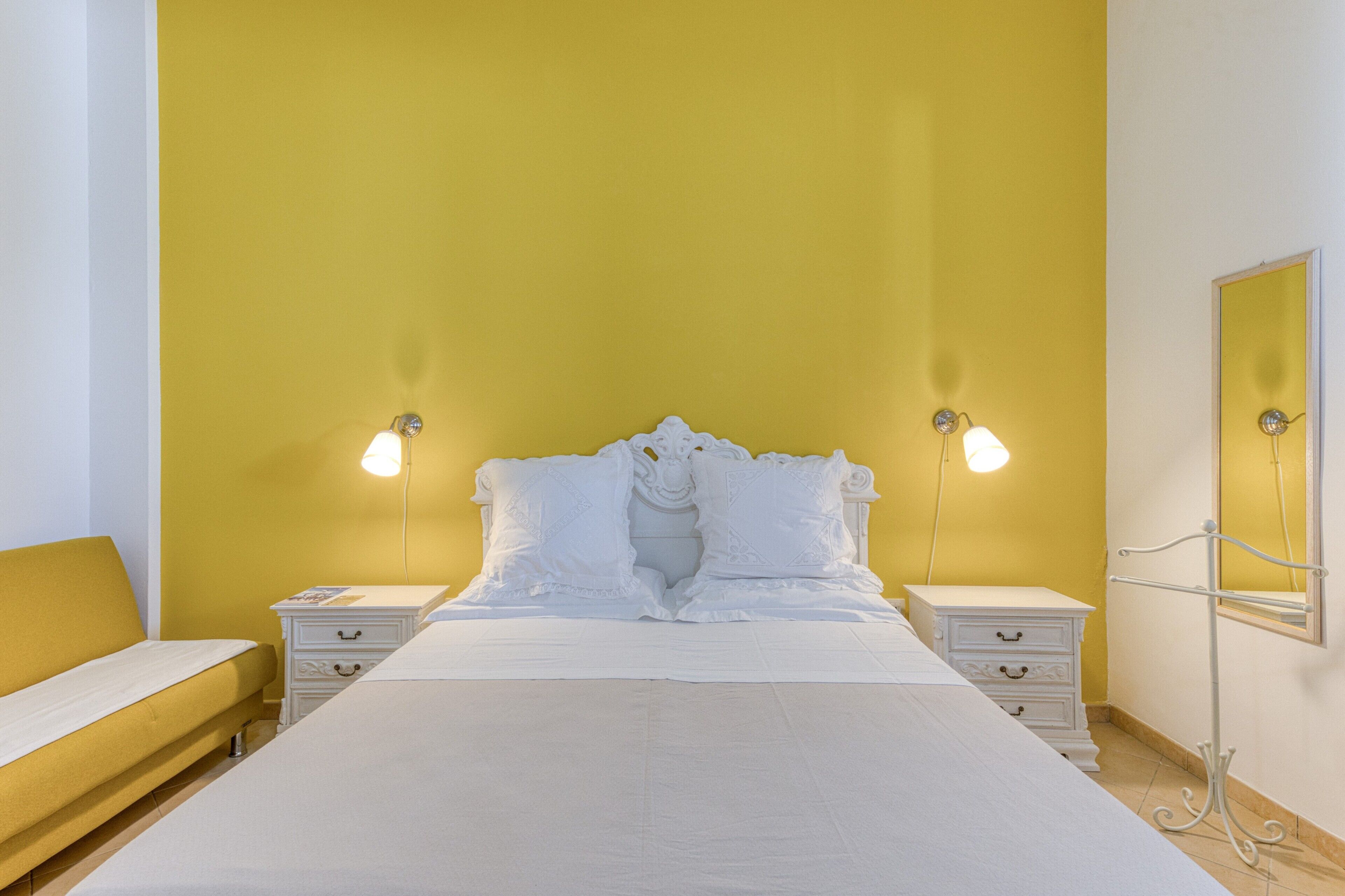 A bed with white bedding, two bedside tables with lamps, a nightstand, and a mirror on the wall.