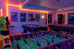 A room with a foosball table, a pool table, and a bar area with neon lights.