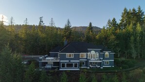 A large, two-story house with a dark roof, multiple windows, and a balcony, surrounded by dense forest. - Mountain Estate | Pickleball, Hot Tub, Game Room (Port Angeles)