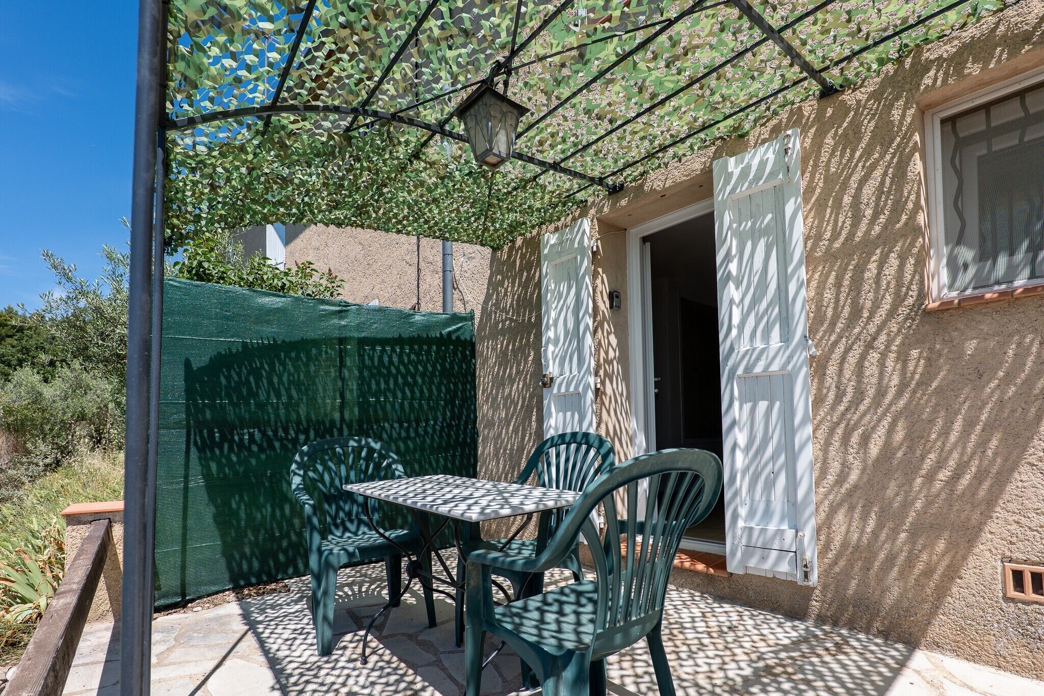 An outdoor patio with a table and chairs, a green mesh canopy, and a building with a textured wall and a window.