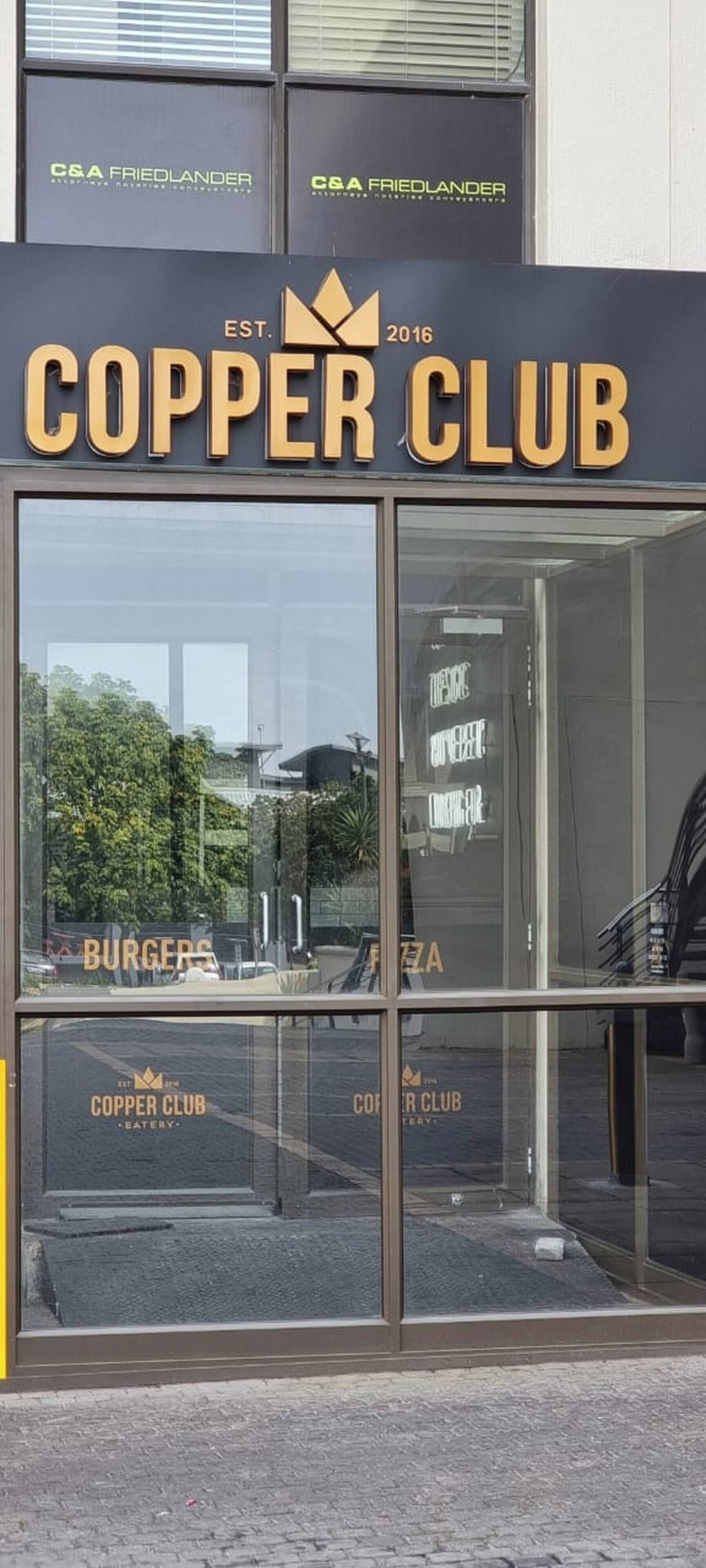 En butiksvindue med et skilt der viser 'COPPER CLUB EST. 2016' og menupunkterne 'BURGERS' og 'PIZZA'.