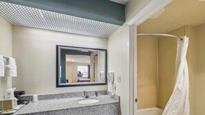Un baño con lavamanos de encimera de granito, un espejo, ducha con cortina blanca y suelo de baldosas.