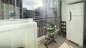 A balcony with a view of buildings, a white refrigerator, and a plant stand. - Home Bikers (São Paulo)