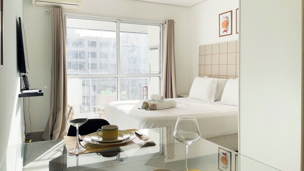 A hotel room with a bed, a bedside table with a wine glass, a TV mounted on the wall, and a window with curtains. - Home Bikers (São Paulo)
