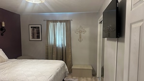 A bedroom with a bed, a bedside table, a television, a cross on the wall, and a window with curtains. - Nice 2-bedroom in charming Smithville near Lake. Onsite boat parking available. (Smithville)