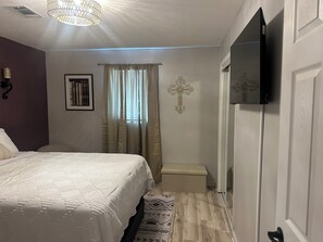 Un dormitorio con cama, mesita de noche, televisor, una cruz en la pared y ventana con cortinas.