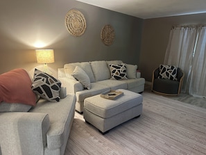 Una sala de estar con un sofá gris, un reposapiés y un sillón, una lámpara y decoración en la pared.