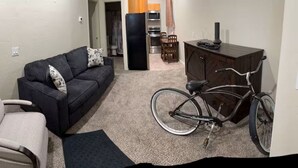 Un salon avec un canapé, un fauteuil, un vélo et, au fond, une petite cuisine.
