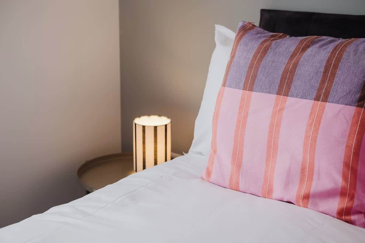 A bed with a white sheet and a striped pillow, a bedside lamp, and a plain wall.